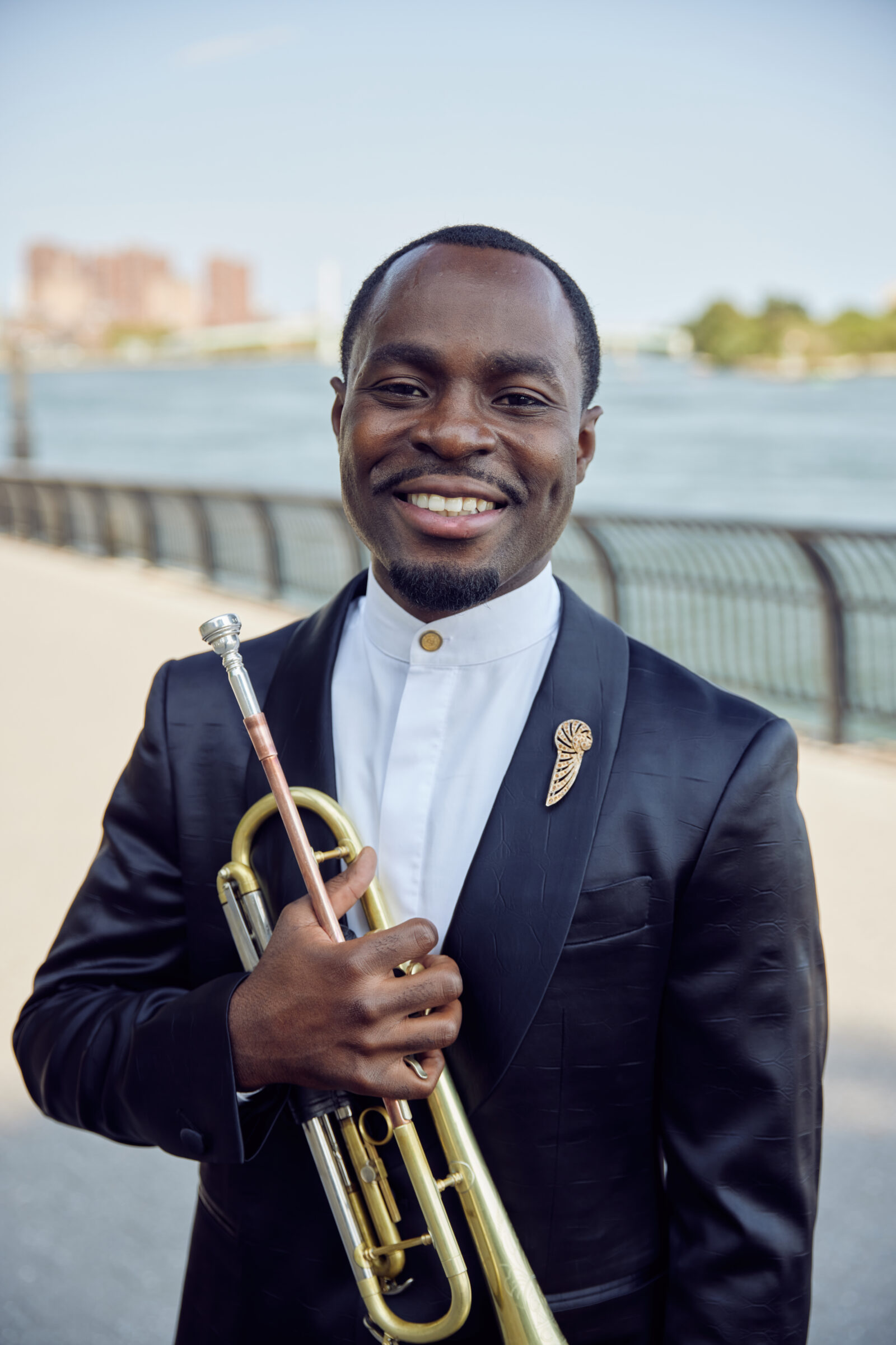 A smiling man in a tuxedo holding a trumpet by the waterfront.